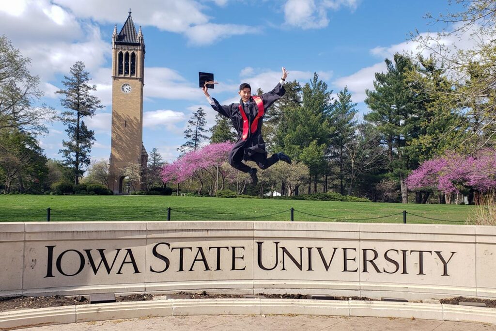 Tan Nguyen, dressed in graduation cap and gown, jumping up in the air above the Iowa State University sign on central campus.