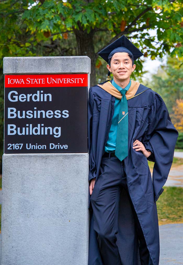 Tan Nguyen, dressed in his master's graduation cap and gown, standing near Gerdin Business Building sign on the central Iowa State university campus.