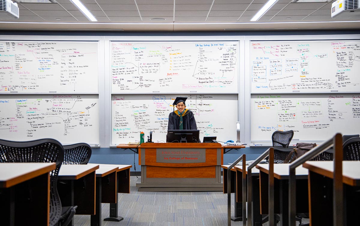 Tan Nguyen, dressed in graduation cap and gown, standing in frontof three large classroom white boards filled with accounting equations.